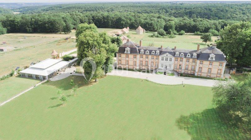 Vue aérienne d'un château entouré de vastes pelouses et de forêts, avec une tente blanche sur la gauche.
