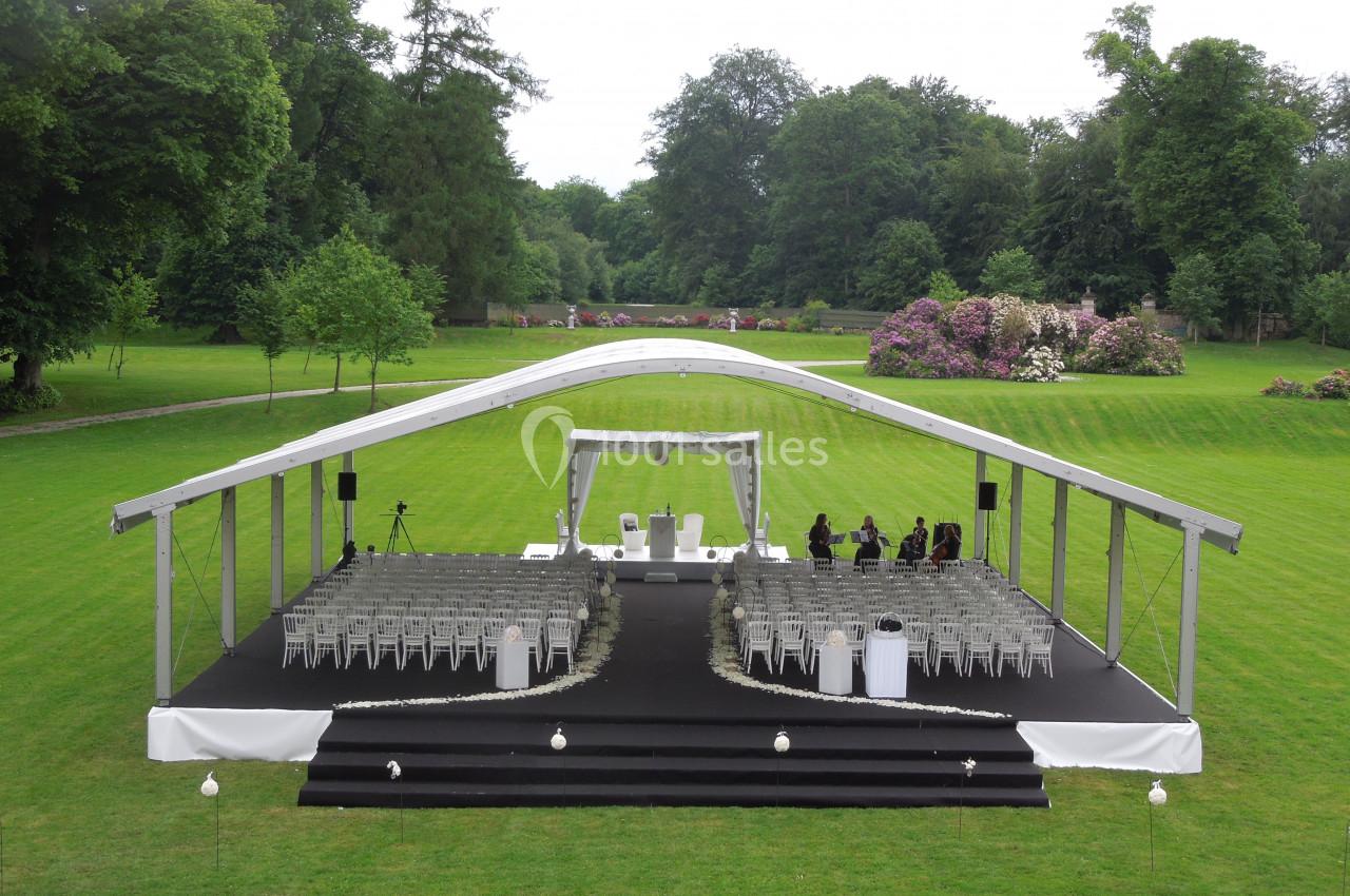 Structure de cérémonie en plein air avec chaises blanches alignées, située sur une pelouse entourée d'arbres et de fleurs.