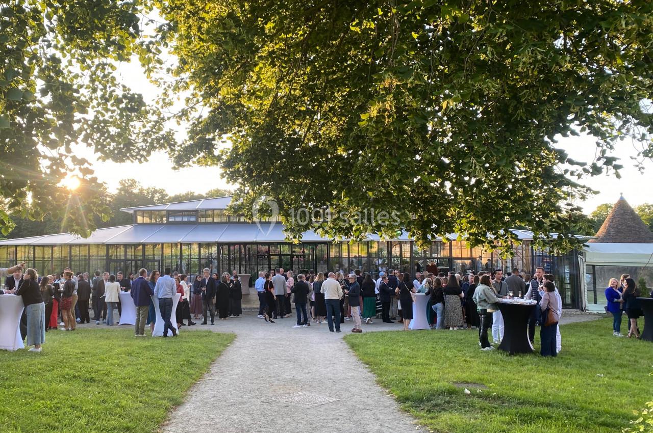 Groupe de personnes rassemblées dans un jardin devant un bâtiment vitré, sous des arbres au coucher du soleil.