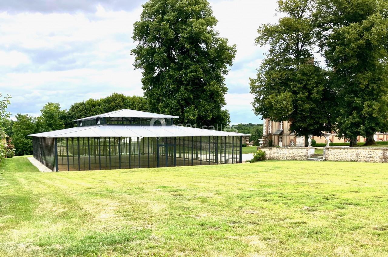 Serre moderne en verre entourée de pelouses et d'arbres, avec un bâtiment ancien en arrière-plan.