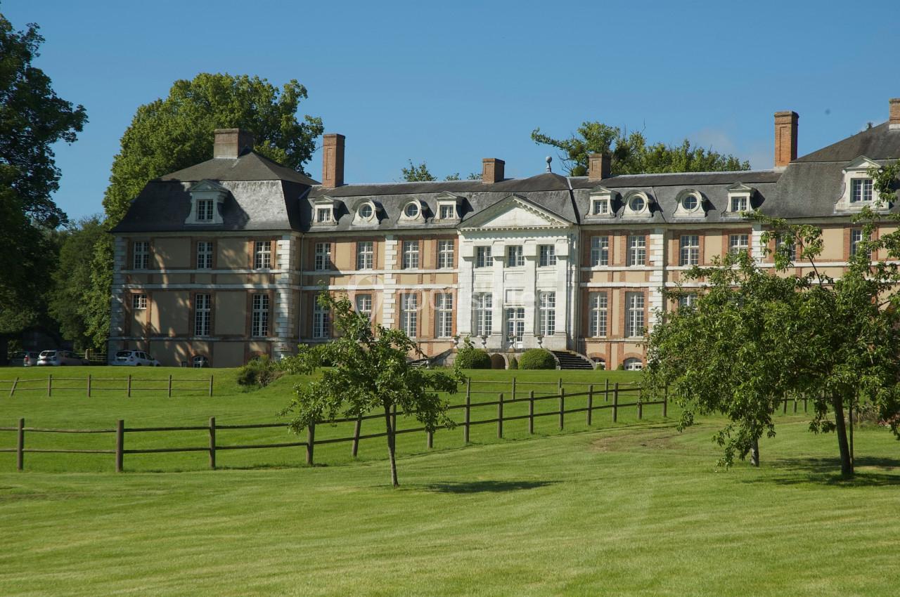 Façade d'un château historique entouré d'un parc verdoyant avec arbres et clôtures en bois.