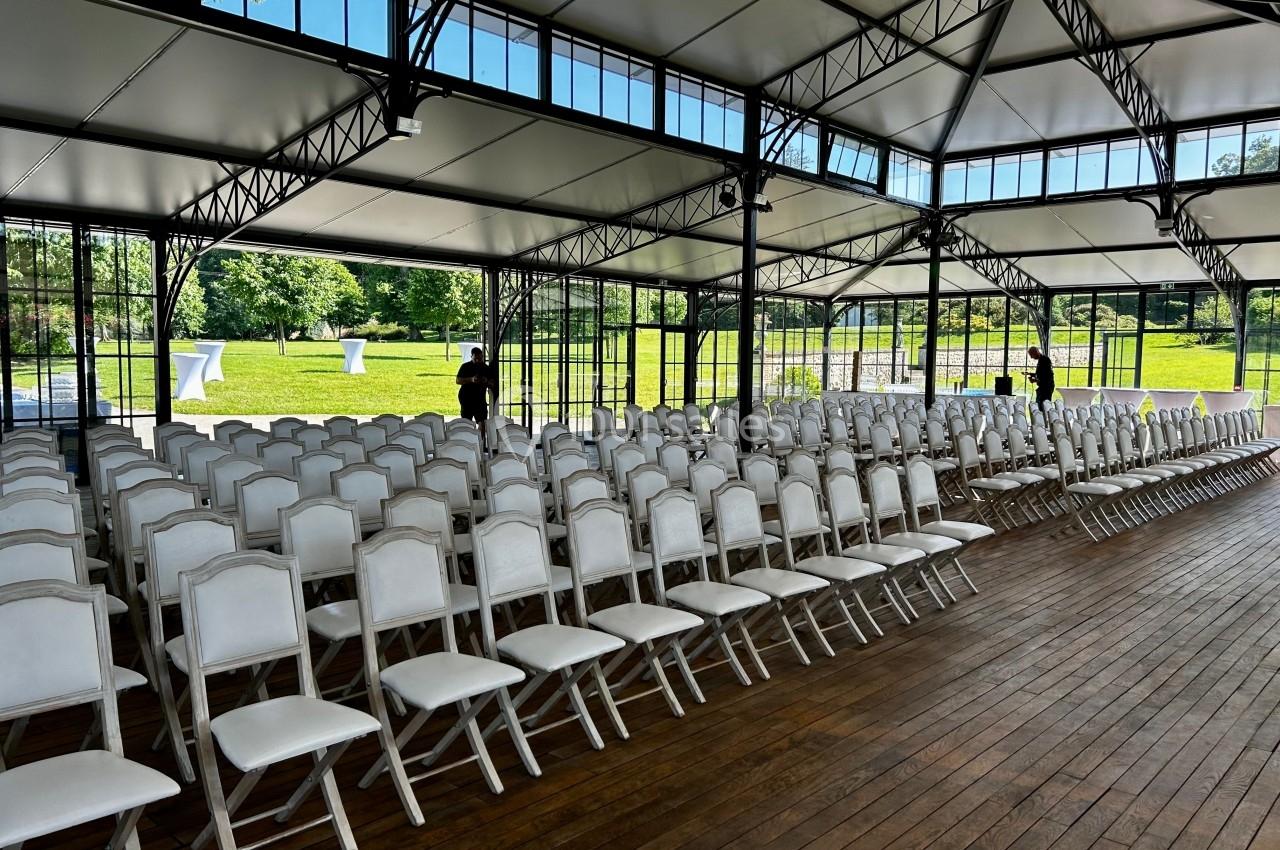Salle lumineuse avec des rangées de chaises blanches alignées sur un sol en bois, ouverte sur un espace vert extérieur.