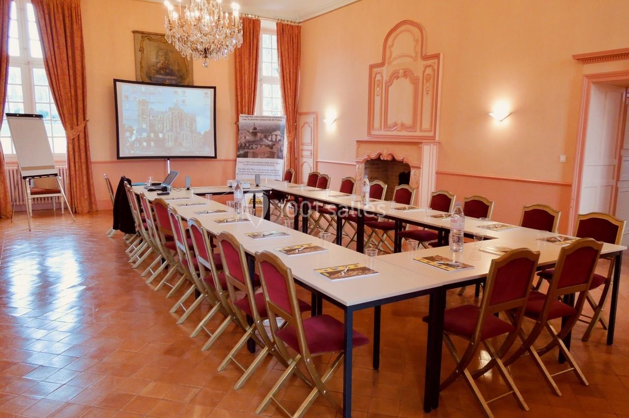 Salle de réunion lumineuse avec tables disposées en U, chaises rouges, écran de projection et décorations classiques.