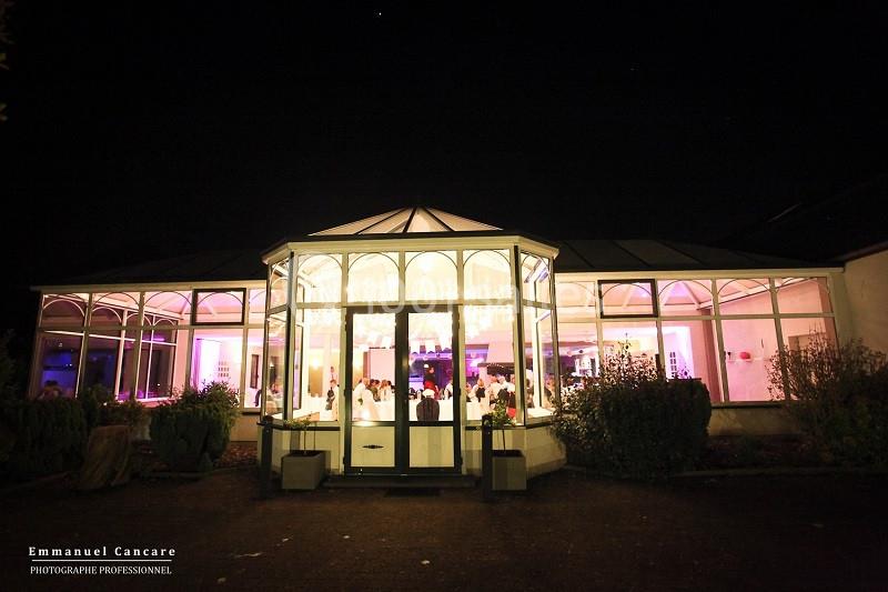 Vue nocturne d'une salle de réception éclairée, avec des baies vitrées laissant entrevoir des invités à l'intérieur.