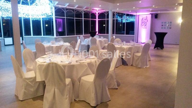Salle de réception lumineuse avec tables rondes dressées, chaises couvertes de housses blanches et éclairage tamisé.