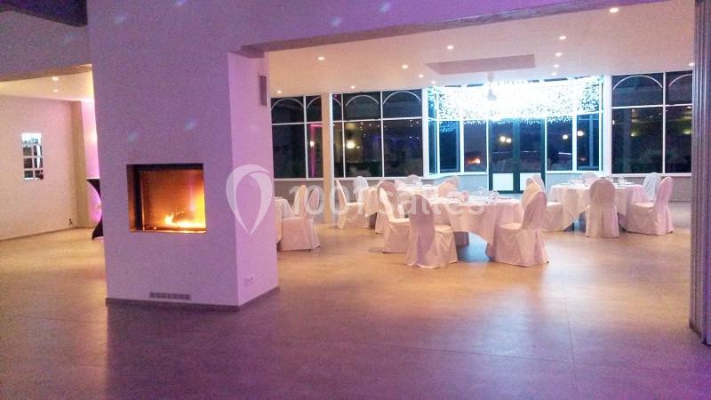 Salle de réception lumineuse avec tables dressées et chaises blanches, cheminée allumée à gauche.
