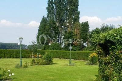 Jardin verdoyant avec pelouse, parterres fleuris, lampadaires verts et haies, entouré d'arbres sous un ciel dégagé.