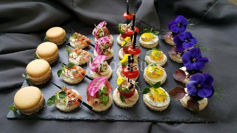 Plateau de bouchées apéritives variées avec macarons salés, canapés garnis et fleurs comestibles sur ardoise noire.