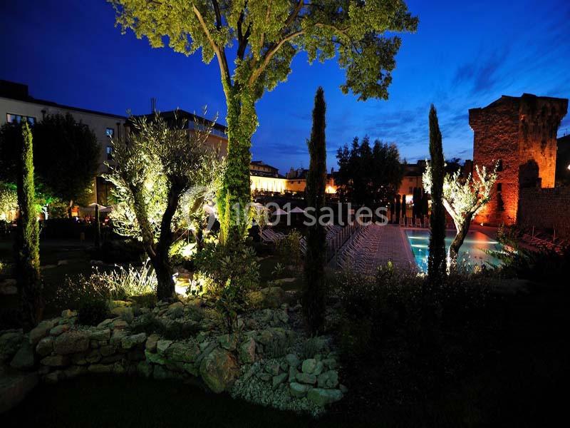 Jardin éclairé de nuit avec piscine, arbres illuminés et bâtiments en arrière-plan sous un ciel bleu foncé.