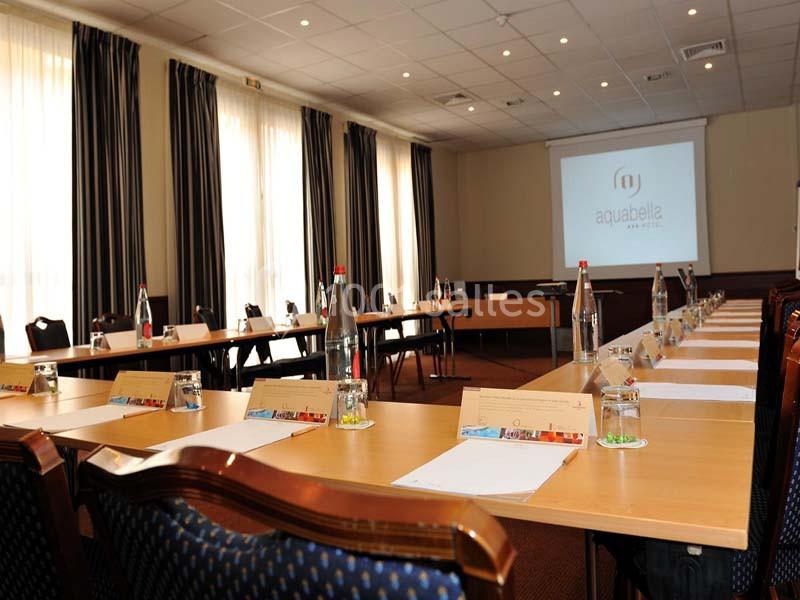 Salle de réunion lumineuse avec une grande table en U, des chaises, des bouteilles d'eau et un écran de projection.
