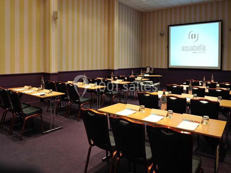 Salle de réunion équipée de tables, chaises, bouteilles d'eau, bloc-notes et écran de projection allumé.