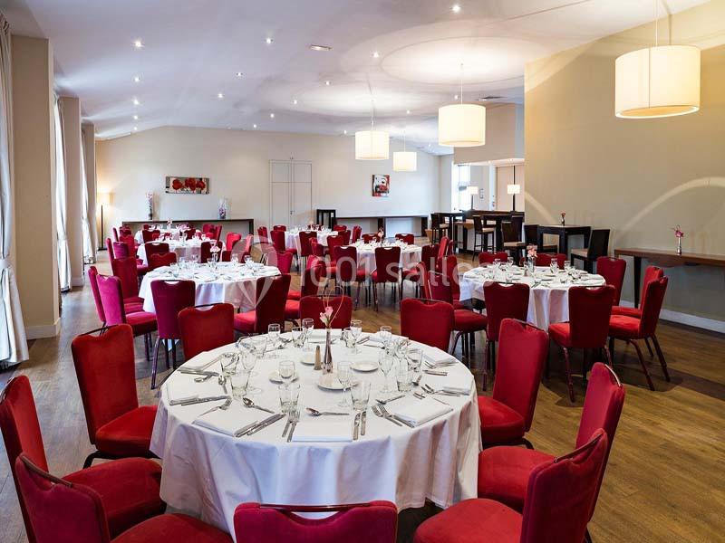 Salle de réception lumineuse avec tables rondes dressées, nappes blanches et chaises rouges.