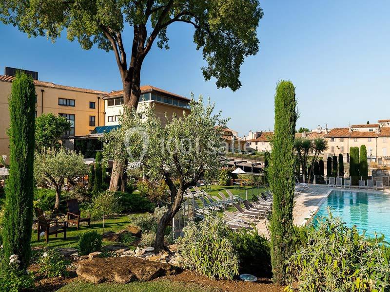 Jardin aménagé avec piscine, chaises longues, arbres et arbustes, entouré de bâtiments en arrière-plan.