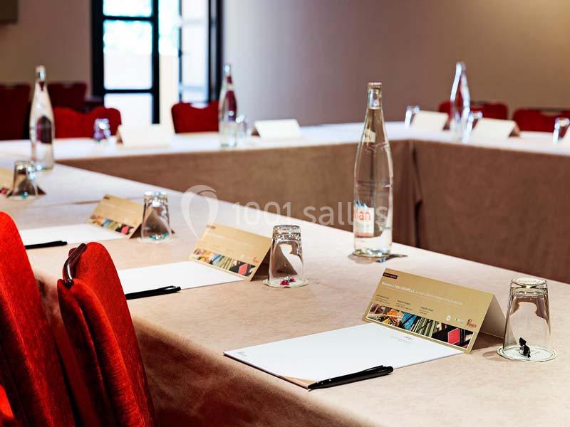 Salle de réunion avec tables couvertes de nappes, disposées en U, équipées de blocs-notes, stylos et bouteilles d'eau.