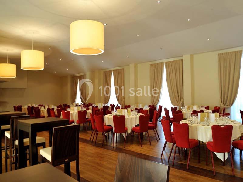 Salle de réception lumineuse avec tables rondes dressées, chaises rouges et grandes fenêtres encadrées de rideaux beiges.