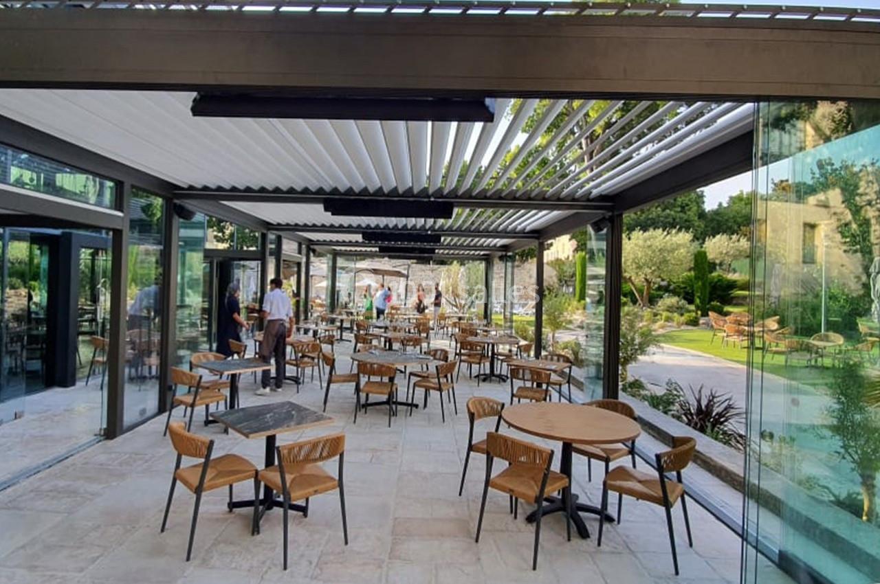 Salle de restaurant lumineuse avec grandes baies vitrées, mobilier en bois et vue sur un jardin extérieur.