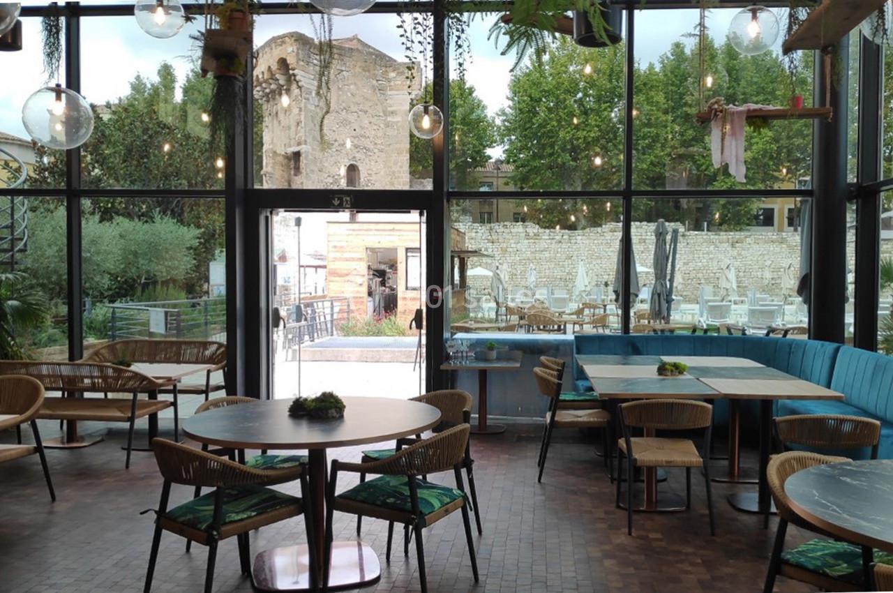 Intérieur d'un café avec des tables en bois, grandes baies vitrées et vue sur une terrasse et des ruines historiques.