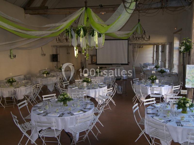 Salle décorée pour un événement avec des tables rondes dressées, chaises blanches et guirlandes vertes suspendues.