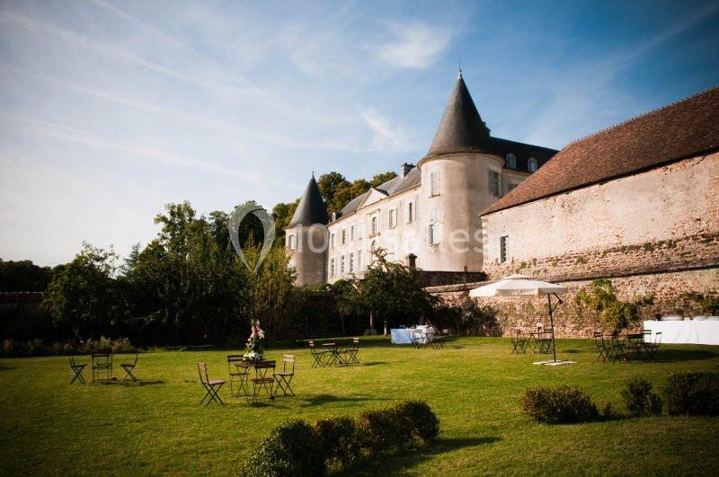 Château ancien avec tours, grand jardin verdoyant, tables et chaises disposées en extérieur sous un ciel dégagé.