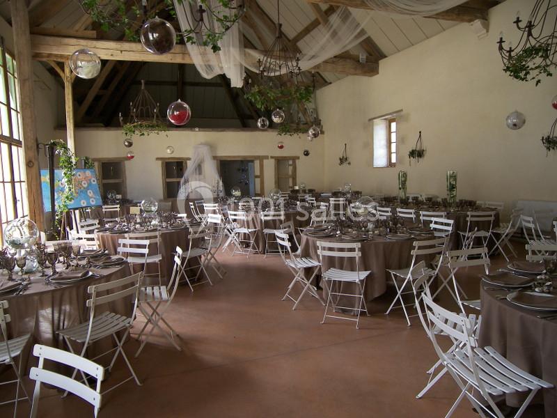 Salle de réception décorée avec des tables rondes, nappes marron, chaises blanches et suspensions florales.