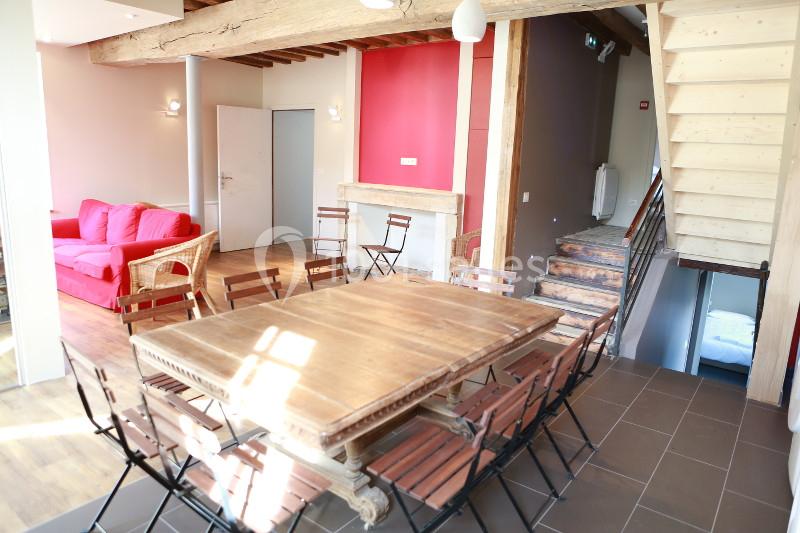 Salle lumineuse avec grande table en bois, chaises, canapé rouge, escalier en bois et mur rouge en arrière-plan.