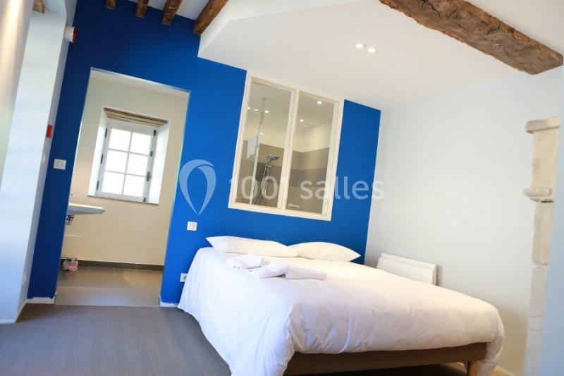 Chambre lumineuse avec lit double, mur bleu, poutres apparentes et salle de bain attenante visible à travers une vitre.