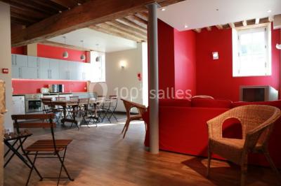 Salle lumineuse avec cuisine ouverte, murs rouges, poutres apparentes, mobilier en bois et fauteuils en osier.