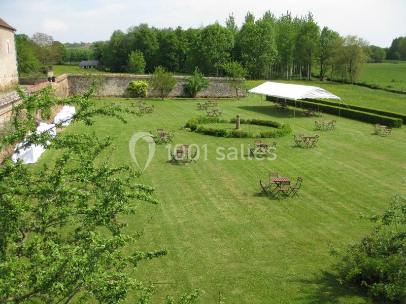 Jardin verdoyant avec tables et chaises espacées, entouré de murs en pierre et d'arbres, avec une tente blanche à droite.