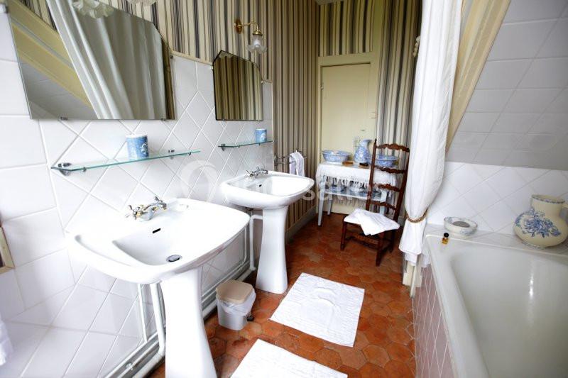 Salle de bain avec deux lavabos, miroirs, baignoire, carrelage blanc et sol en terre cuite.