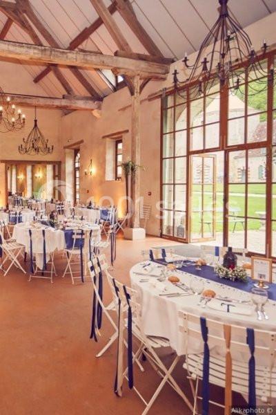 Salle de réception lumineuse avec tables rondes dressées, chaises blanches et rubans bleus, grandes fenêtres en arrière-plan.