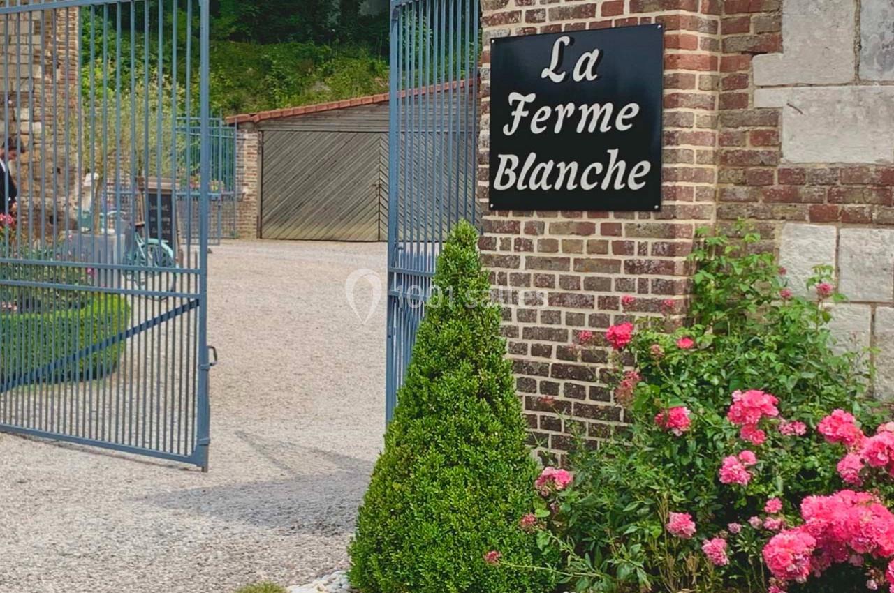 Entrée d'une ferme avec portail ouvert, panneau ’La Ferme Blanche’, buisson taillé et rosiers en fleurs.