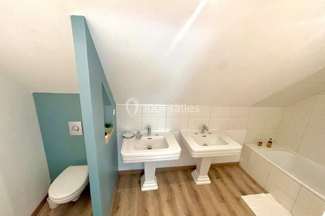 Salle de bain mansardée avec deux lavabos, une baignoire, un WC séparé par une cloison et un sol en parquet.