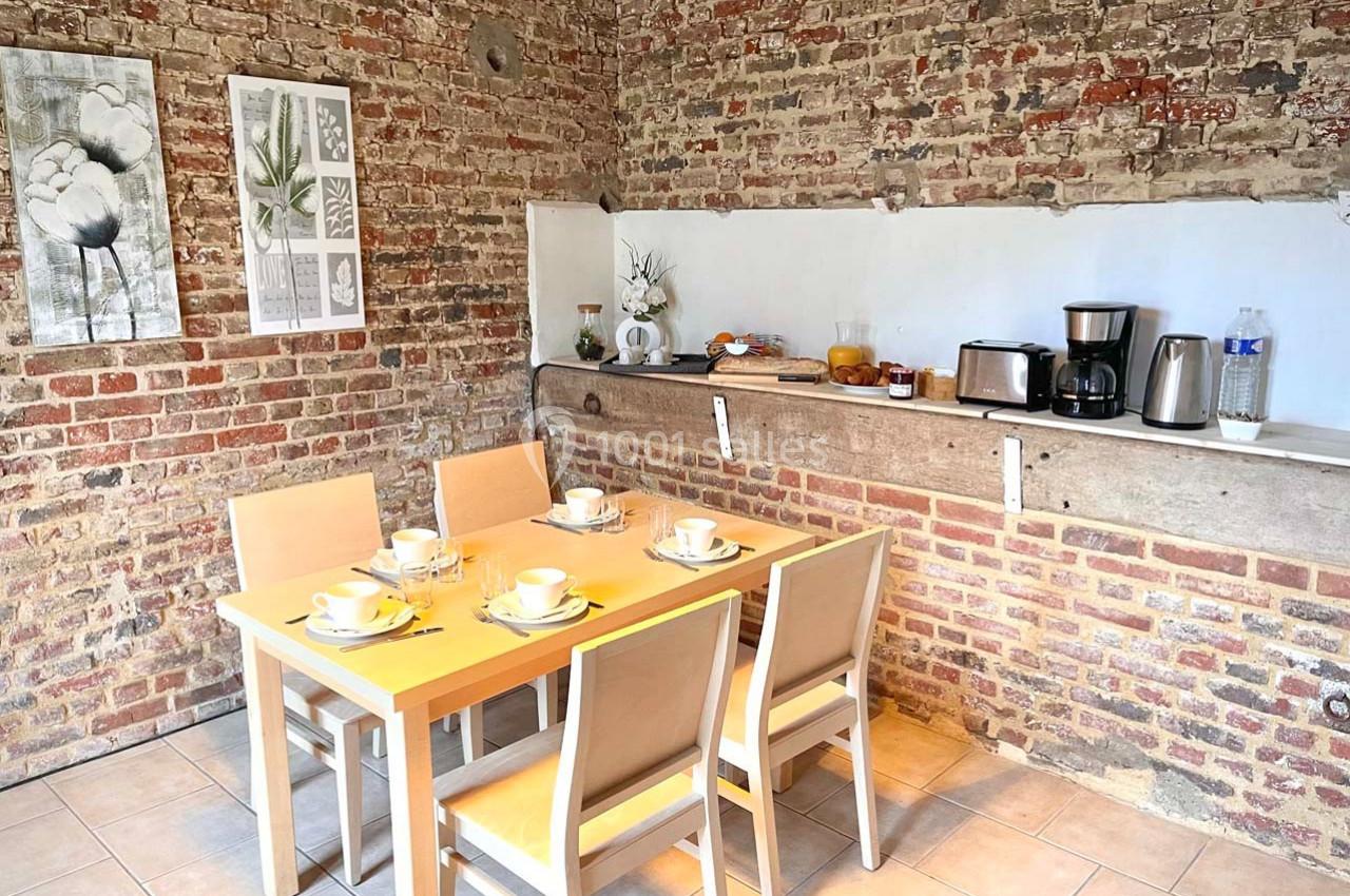 Salle à manger avec table en bois, chaises, vaisselle disposée et mur en briques apparentes.