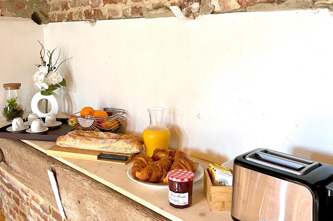 Petit-déjeuner avec croissants, baguette, confiture, jus d'orange, fruits, cafetière et grille-pain sur un buffet en bois.