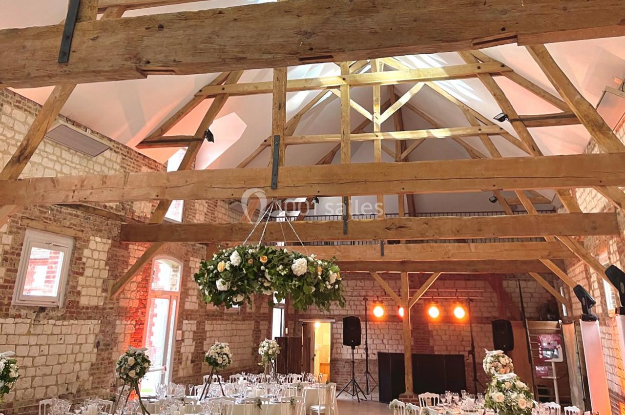 Salle de réception avec poutres apparentes, murs en pierre, tables dressées et décorations florales suspendues.