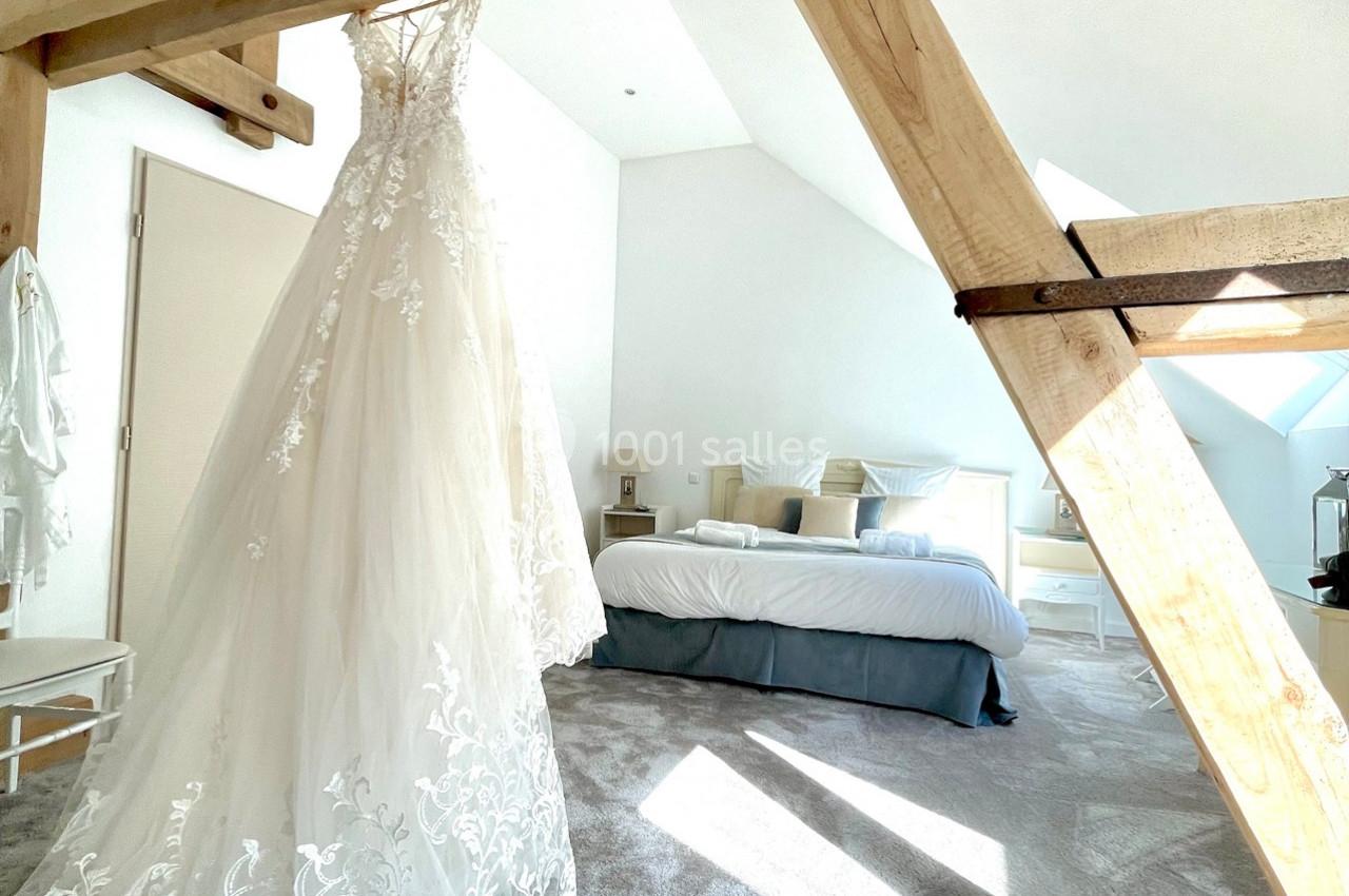 Robe de mariée blanche suspendue dans une chambre lumineuse avec lit double et poutres apparentes.
