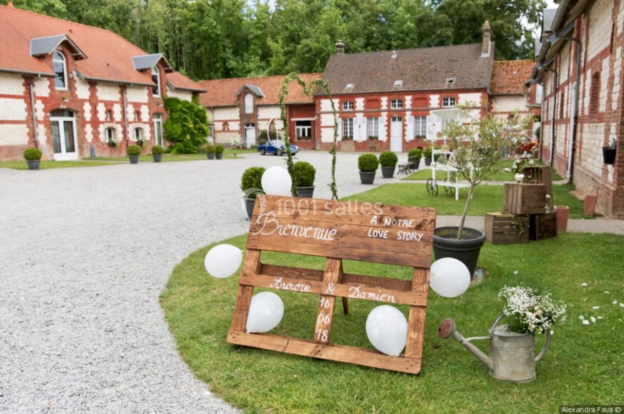 Panneau en bois décoratif avec ballons blancs, placé dans une cour devant des bâtiments en briques et un jardin fleuri.