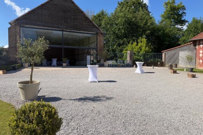 Miniature Location salle Épagne-Épagnette (Somme) - La Ferme Blanche #33 Cour extérieure en gravier avec une grange ouverte, des tables hautes blanches et des plantes en pot, entourée de verdure.