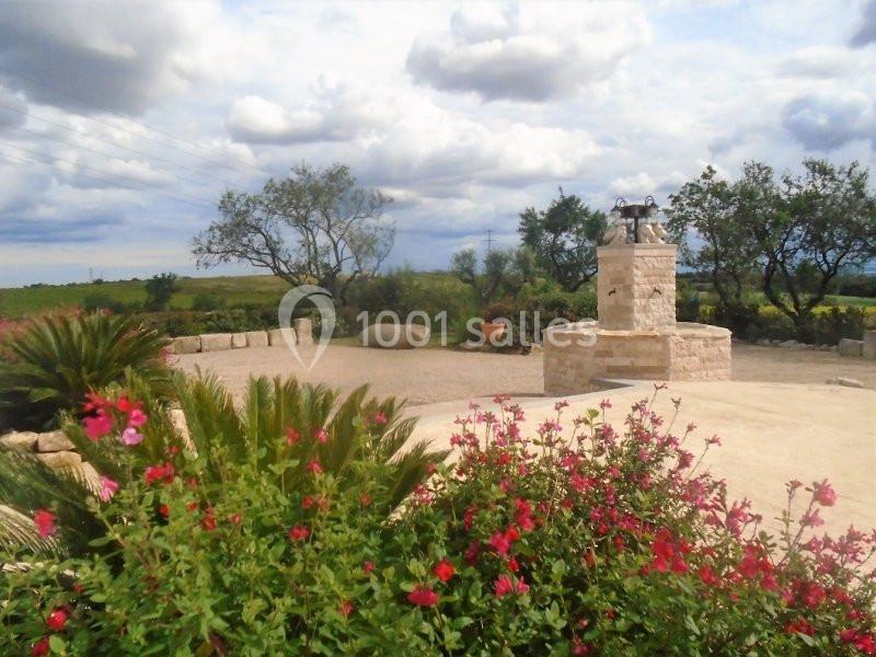 Jardin méditerranéen avec fleurs rouges au premier plan, un puits en pierre et des arbres sous un ciel partiellement nuageux.