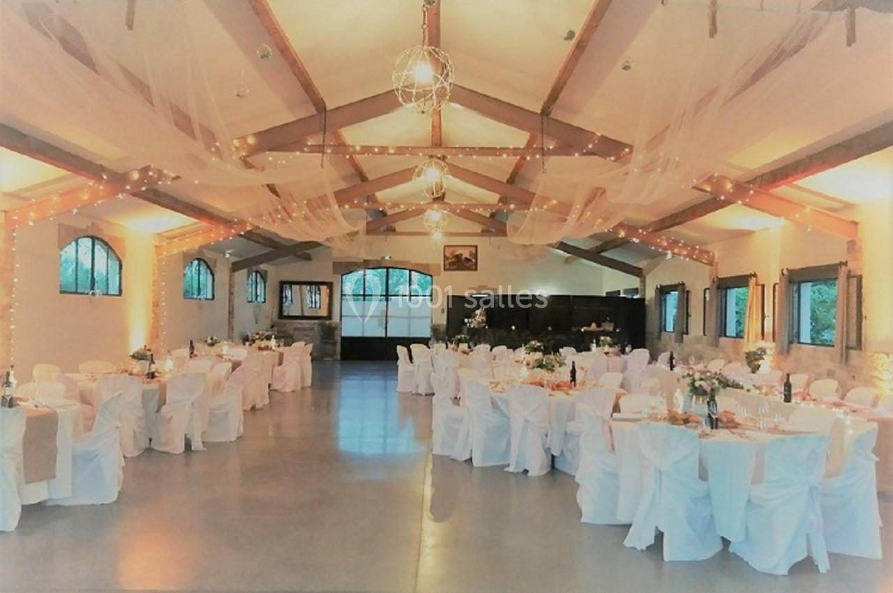 Salle de réception décorée avec des tables rondes, nappes blanches, guirlandes lumineuses et voilages suspendus.