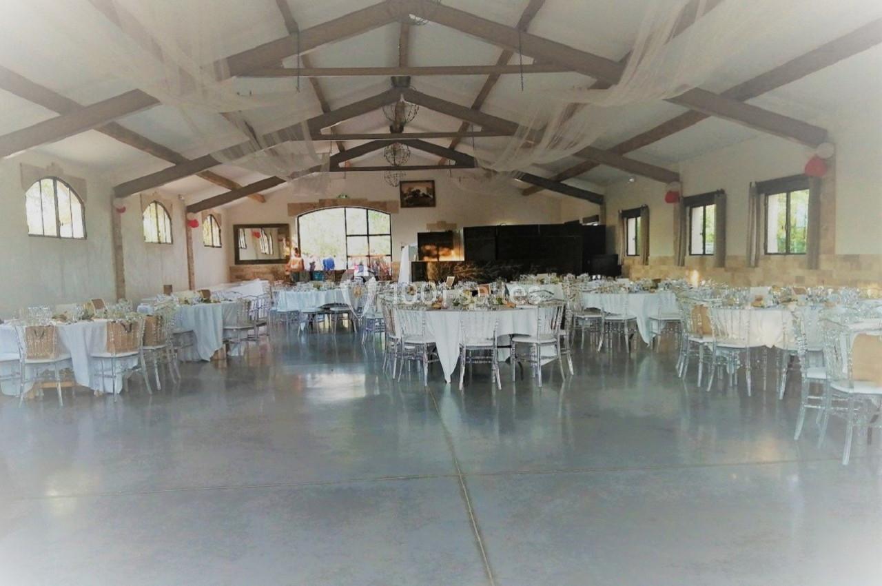 Salle de réception décorée avec des tables rondes et rectangulaires dressées, sous une charpente en bois apparente.