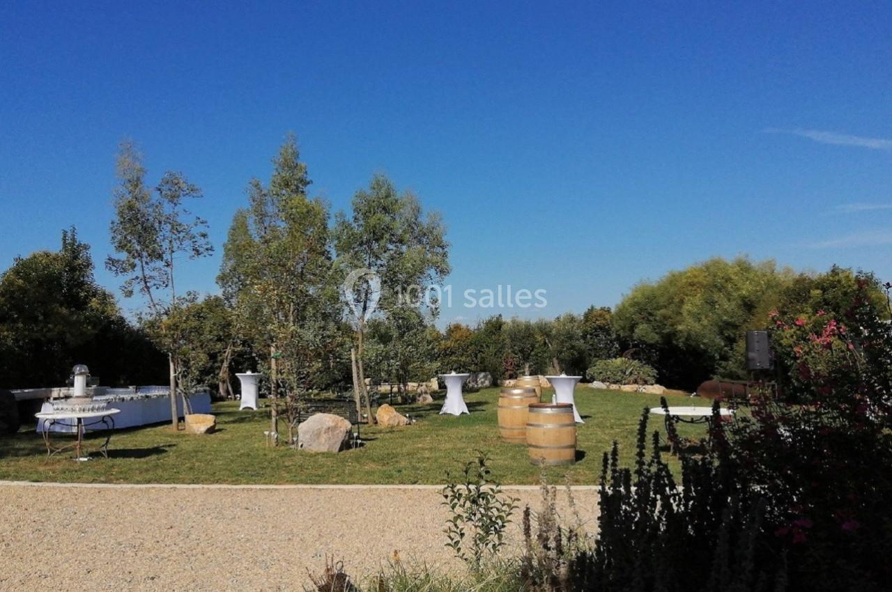 Espace extérieur avec pelouse, tables hautes, tonneaux en bois et arbres sous un ciel bleu dégagé.