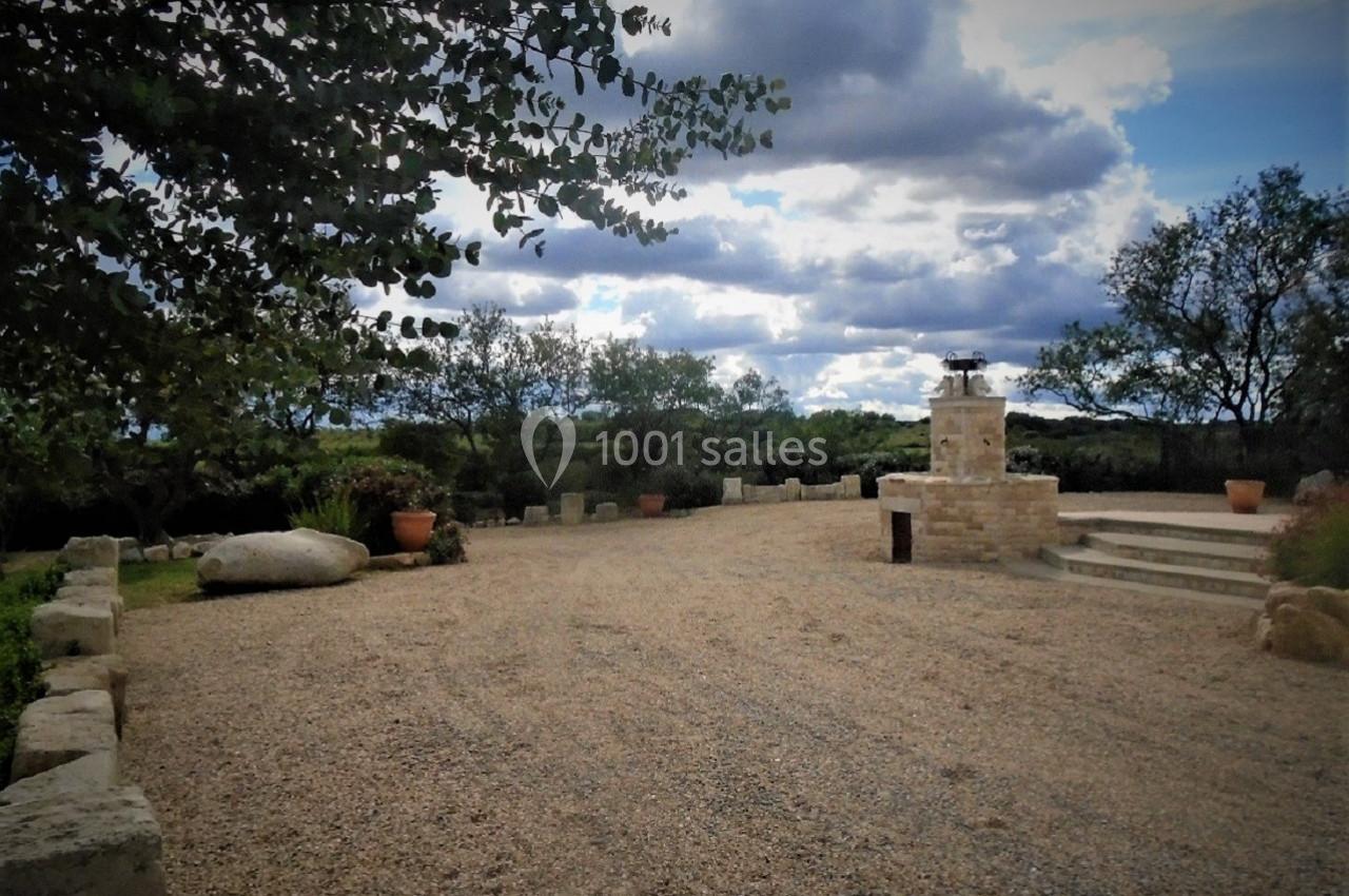Cour extérieure en gravier avec un barbecue en pierre, entourée d'arbres et de pots de fleurs sous un ciel nuageux.