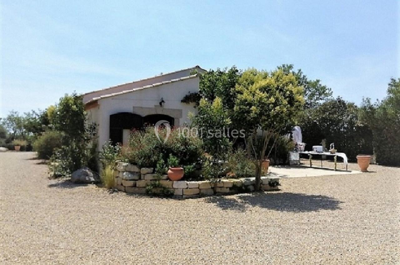 Petite maison entourée de végétation et d'une terrasse aménagée avec des tables sur un sol gravillonné.