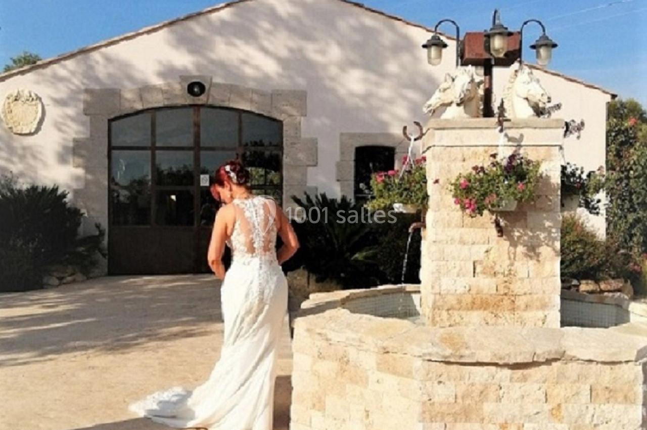 Une mariée en robe blanche marche près d'une fontaine en pierre devant un bâtiment lumineux sous un ciel bleu.