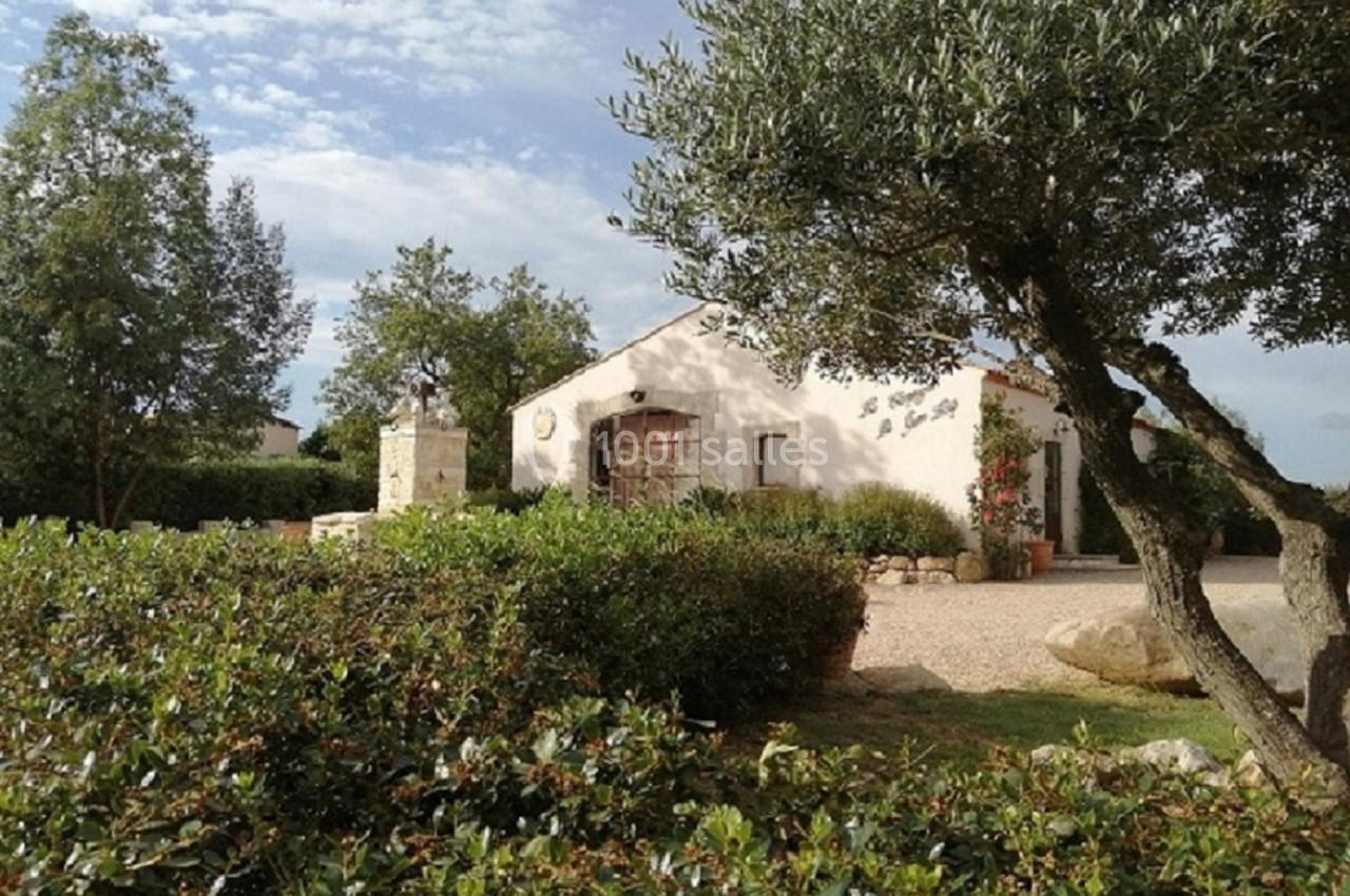 Maison blanche entourée de végétation méditerranéenne, avec un arbre au premier plan et un ciel partiellement nuageux.