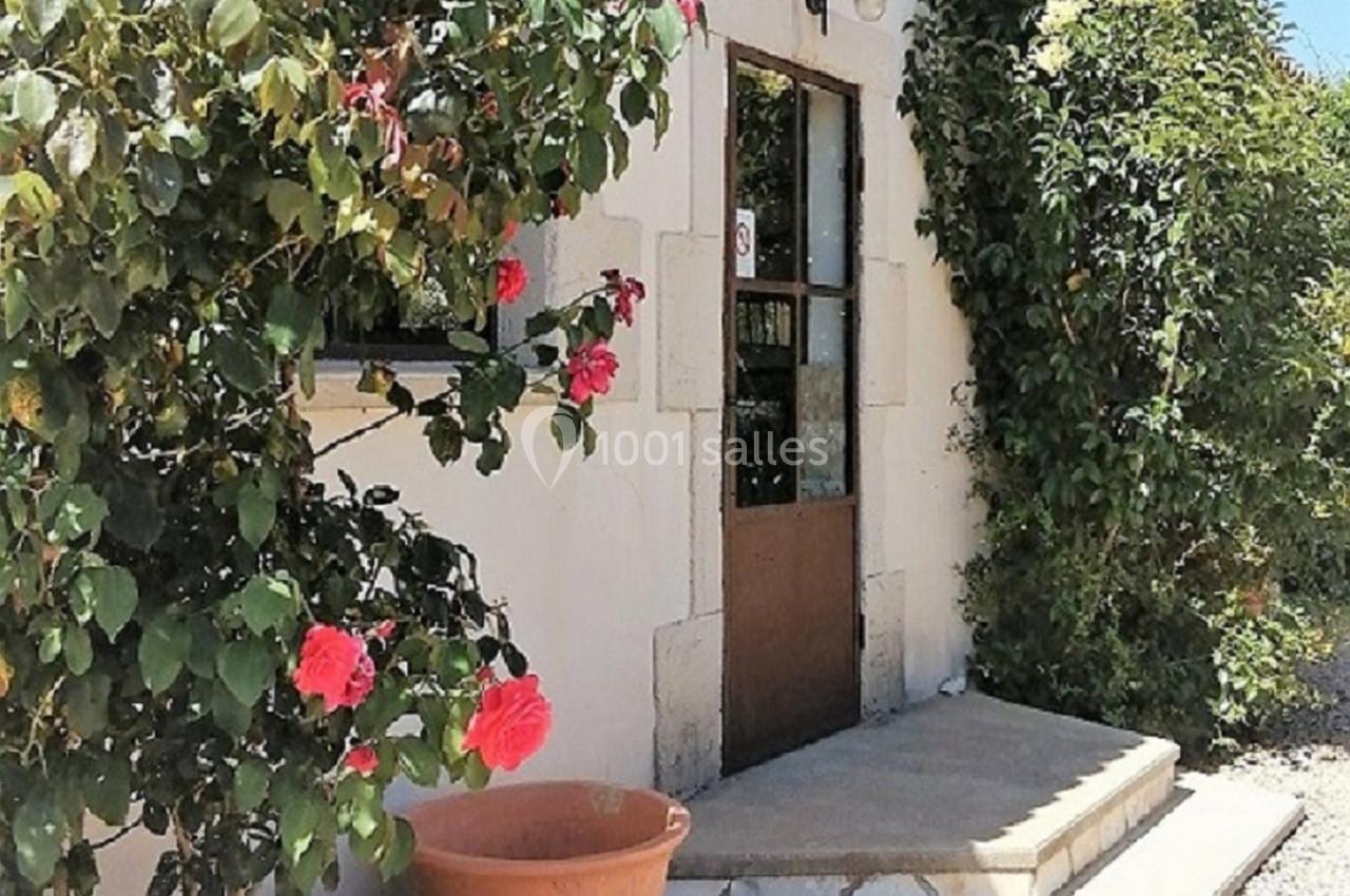 Façade d'une petite maison en pierre avec porte en bois, entourée de rosiers et d'un pot en terre cuite.