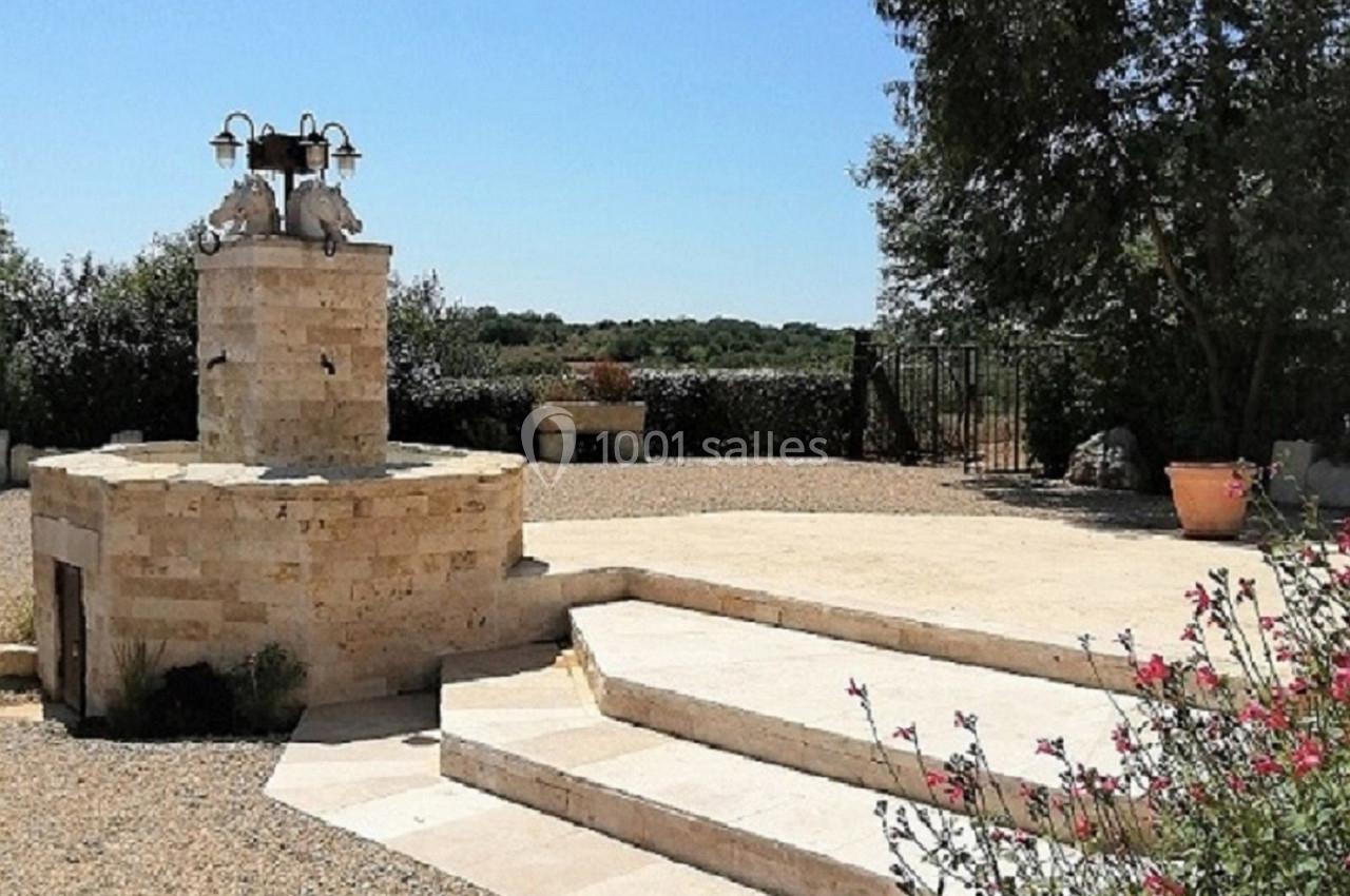 Cour extérieure ensoleillée avec un petit puits en pierre, des escaliers, des plantes et des arbres en arrière-plan.