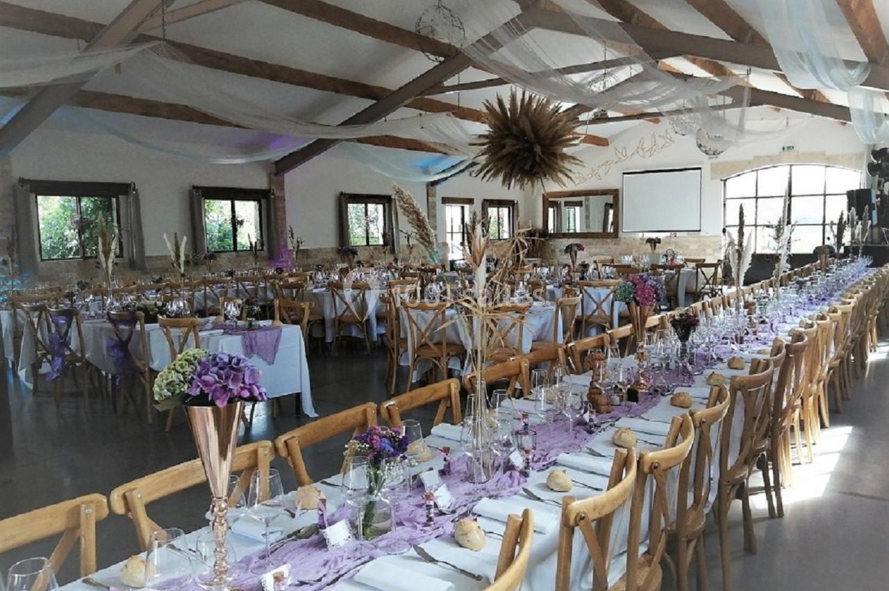Salle de réception décorée pour un événement, avec tables alignées, nappes violettes, fleurs et chaises en bois.