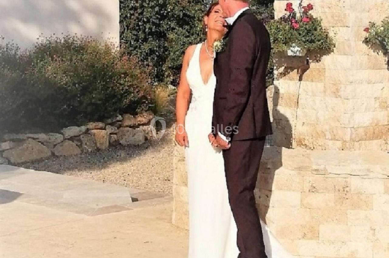 Un couple en tenue de mariage pose près d'une fontaine en pierre devant un bâtiment blanc.